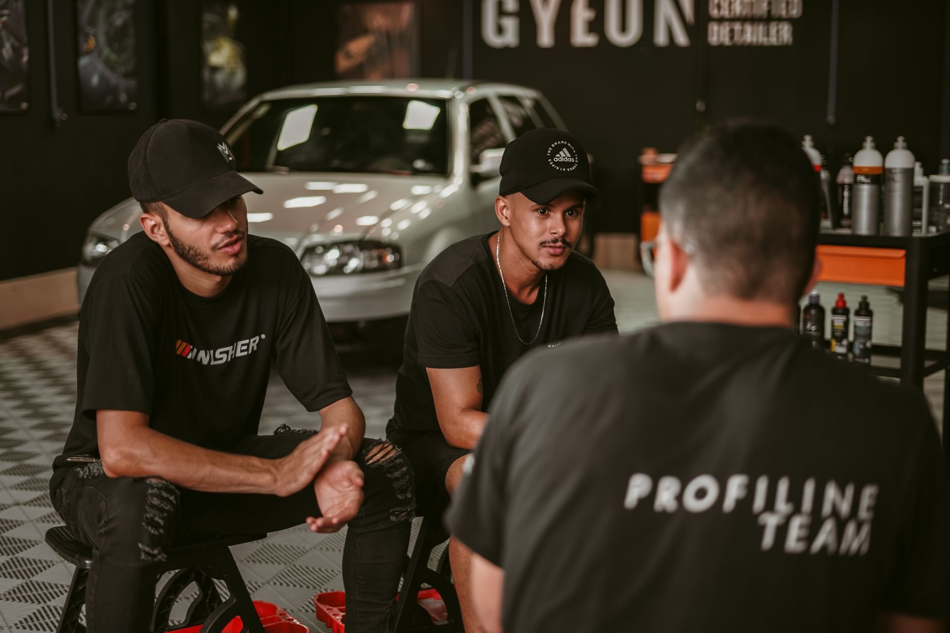Team of men discussing in a garage with car detailing products and vehicle in the background.